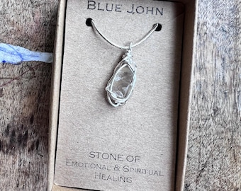 May include: A clear quartz pendant necklace with a silver-colored wire wrap, displayed in a small cardboard box. The box has the text "BLUE JOHN" at the top and "STONE OF EMOTIONAL & SPIRITUAL HEALING" at the bottom. The necklace is on a wooden surface.