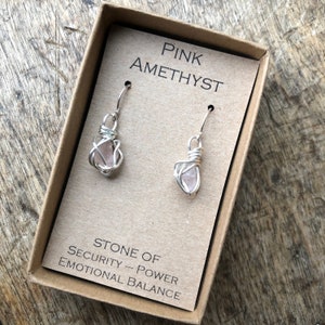 May include: Pink Amethyst earrings in a small box. The earrings are made with wire wrapped around small pink amethyst stones. The box has the text "PINK AMETHYST" and "STONE OF SECURITY - POWER EMOTIONAL BALANCE" printed on it.