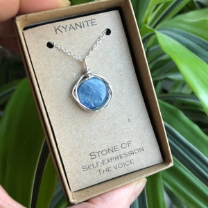 May include: A silver necklace with a blue kyanite stone pendant. The pendant is wrapped in silver wire and has a round shape. The necklace is in a small box with the words "Kyanite" and "Stone of Self-Expression The Voice" printed on it.