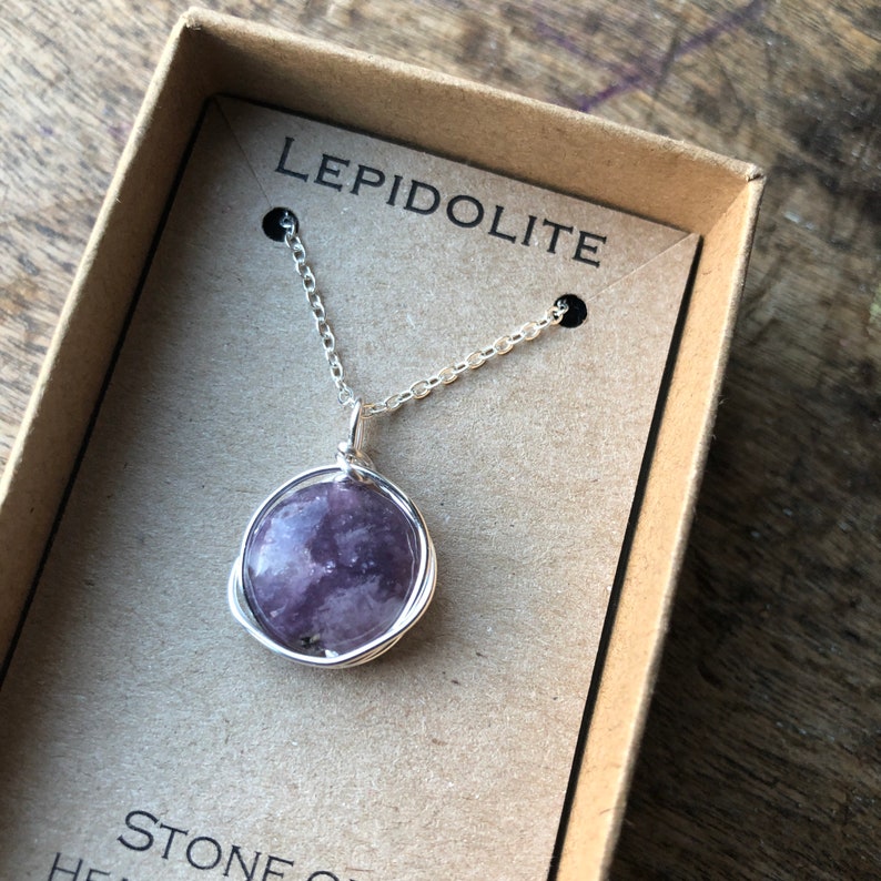 Pu&ograve; includere: Una collana a catena d'argento con un ciondolo in pietra di lepidolita viola avvolto in filo d'argento. La collana &egrave; in una scatola marrone con le parole "Lepidolite" e "Pietra" stampate su di essa.