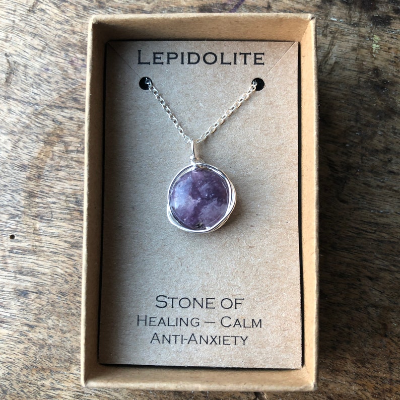 Pu&ograve; includere: Una collana con catena d'argento con un ciondolo in pietra di Lepidolite viola in una piccola scatola. La scatola ha il testo "LEPIDOLITE" e "PIETRA DI GUARIGIONE - CALMA ANTI-ANSIA" stampato su di essa.