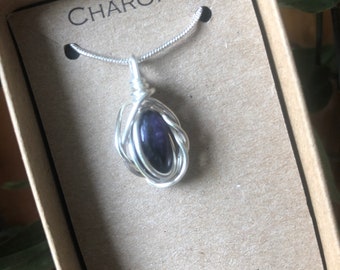 Könnte beinhalten: Eine silberne Kettenkette mit einem Anhänger aus lila Charoit-Stein, der in Silberdraht gewickelt ist. Die Kette befindet sich in einer Schachtel mit dem aufgedruckten Wort "CHAROITE". Die Schachtel trägt auch den Schriftzug "STEIN DER ENERGIE".