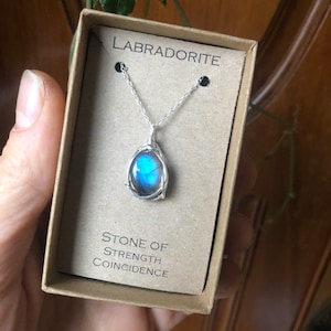 May include: A silver chain necklace with a blue labradorite gemstone pendant. The pendant is wrapped in silver wire and the box says "Labradorite Stone of Strength Coincidence".