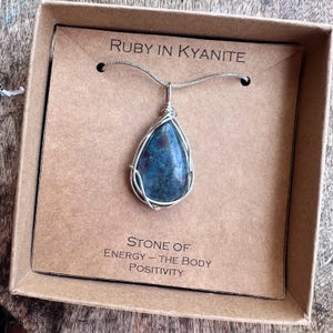May include: A silver necklace with a teardrop-shaped pendant. The pendant features a blue and red stone encased in a silver wire frame. The necklace is displayed in a brown cardboard box. Text on the box reads "Ruby in Kyanite" and "Stone of Energy - The Body Positivity."