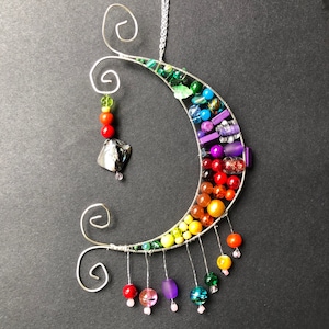 Può includere: Una scultura in filo metallico a forma di mezzaluna decorata con perline colorate in un motivo arcobaleno. Le perline sono infilate sul filo e pendono dalla parte inferiore della scultura.