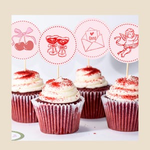 May include: Four red velvet cupcakes with white frosting and red sprinkles. Each cupcake has a decorative topper with a Valentine's Day theme, including cherries, champagne glasses, an envelope with a heart, and a cupid.