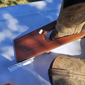 May include: A wooden boot jack with a curved design, made of dark brown wood. A pair of brown cowboy boots with decorative stitching are placed on the jack. The text "mahogany Boot Jack" is visible on a tag.