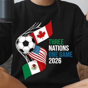 May include: Black sweatshirt featuring a soccer ball and the flags of Canada, the United States, and Mexico. Text reads "THREE NATIONS ONE GAME 2026". The design is in white, green, red, and blue.