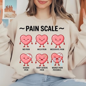 May include: Beige sweatshirt with a graphic of a pain scale featuring cartoon hearts. The scale ranges from "No Pain" to "Worst Pain Possible." Each heart has a different expression and some have bandages.