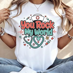 Może przedstawiać: Biała koszulka z kolorową grafiką z napisem "You Rock My World". Projekt zawiera czapkę pielęgniarki, stetoskop, pigułki i strzykawki. Tekst jest czerwony i turkusowy, ze stetoskopem w kształcie serca.