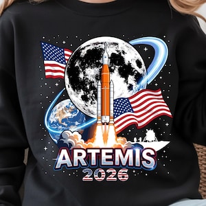 Puede incluir: Sudadera negra con un gráfico de un cohete lanzándose hacia la luna, con la Tierra y banderas americanas de fondo. La palabra "ARTEMIS" y el año "2026" se muestran debajo del cohete.