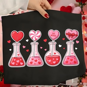 May include: Black tote bag featuring four pink flasks with heart-shaped toppers. The flasks contain pink liquid and heart-shaped designs. One heart-shaped topper says "BE MINE". Small hearts are scattered around the flasks.