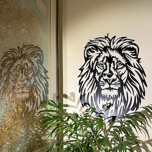 May include: Black and white metal lion head wall art. The lion's face is intricately detailed with a flowing mane. The artwork is mounted on a white wall, with a reflection of a map on the left. Green plants are in the foreground.