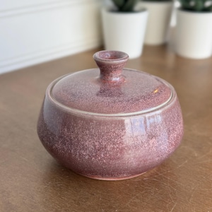 May include: A round ceramic sugar bowl with a lid, featuring a speckled mauve and dusty rose glaze. The bowl has a wide, curved body and a small, knob-shaped handle centered on the lid, showcasing a smooth, glossy finish throughout the piece.