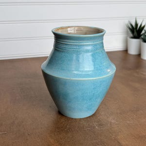 May include: A ceramic vase with a glossy turquoise glaze, featuring a wide, angular body that tapers toward a narrow neck and a flared rim. The interior of the vessel is finished in a smooth, light beige glaze, contrasting with the vibrant blue exterior.