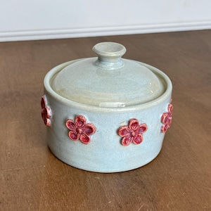 May include: A round, light blue ceramic sugar bowl with a matching lid and a small knob handle. The exterior of the bowl is decorated with several small, raised, five-petaled red flowers evenly spaced around the circumference of the container.