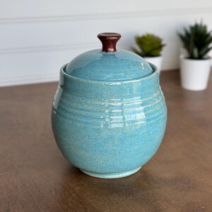 May include: A round, turquoise ceramic jar with a speckled finish and horizontal ribbed detailing. The jar features a matching lid topped with a small, dark brown knob handle. The surface has a glossy glaze with subtle variations in color.