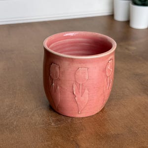 May include: A cylindrical ceramic cup in a solid dusty pink hue, featuring a repeating embossed tulip flower pattern around the exterior. The interior of the vessel shows subtle horizontal ridges from the pottery throwing process.