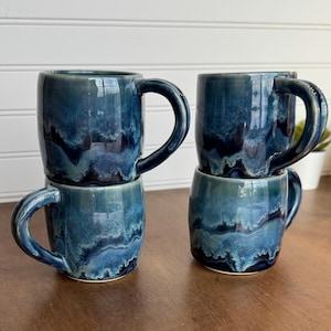 May include: Four stacked ceramic mugs with a glossy, reactive glaze in shades of blue and teal. The mugs have a rounded shape with a curved handle. The glaze creates a marbled effect, adding visual interest to the drinkware.