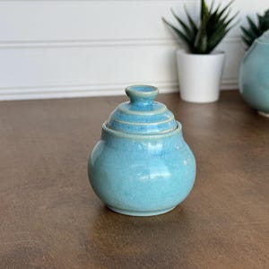 May include: A small, round, light blue ceramic sugar bowl with a matching lid. The bowl features a smooth, glossy glaze with subtle variations in color. The lid has a tiered, stepped design that tapers to a small, circular knob at the top.