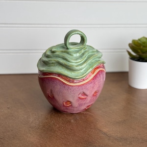 May include: A handmade ceramic jar with a lid. The jar is a vibrant pink with red accents and textured details. The lid is a wavy, light green with a loop handle. The jar is on a wooden surface.