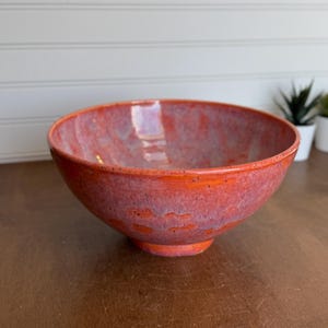 May include: A round ceramic bowl featuring a vibrant glaze with blended shades of bright orange, deep red, and soft purple. The surface has a glossy finish with subtle variations in color intensity throughout the exterior and interior.