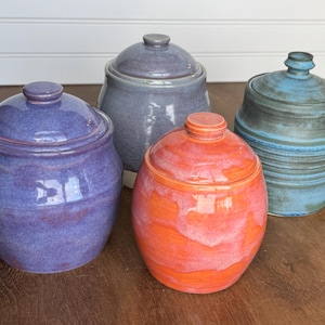 May include: Four ceramic jars with lids in shades of purple, gray, orange, and blue. The jars have a glossy finish and varying shapes, with rounded and cylindrical designs. The jars are arranged on a wooden surface.
