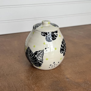 May include: Cream-colored ceramic jar with a lid, adorned with black leaf patterns and small green dots. The jar has a rounded shape and a small knob on the lid. The design is simple and elegant.