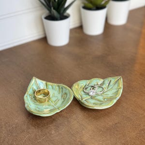 May include: Two small, leaf-shaped ceramic trinket dishes with a glossy, light green glaze and visible veining. One dish holds a plain gold band, while the other holds a silver ring featuring a central stone surrounded by a halo of smaller stones.