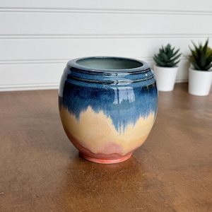 May include: A round ceramic vase featuring a smooth, glossy glaze that transitions from a deep navy blue at the top rim to a warm golden yellow in the center, fading into a soft coral pink at the base with a wide, open circular mouth.