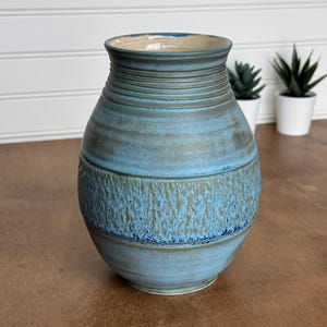 May include: A ceramic vase with a rounded body and a flared rim, featuring a blue glaze with horizontal ridges at the neck and a textured, mottled green and blue band around the center, showcasing a smooth finish on the base and upper sections.