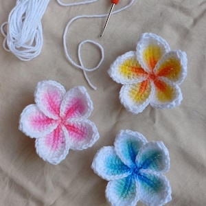 May include: Three crocheted plumeria flowers in white, pink, yellow, orange, and blue hues. A white yarn ball and an orange-handled crochet hook are also visible. The flowers have a detailed, textured appearance, suggesting handmade craftsmanship.