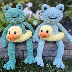 May include: Two crocheted frog plushies, one teal and one light blue, each wearing a yellow and orange duck-shaped floatie. The frogs are sitting on a stone ledge in front of greenery.
