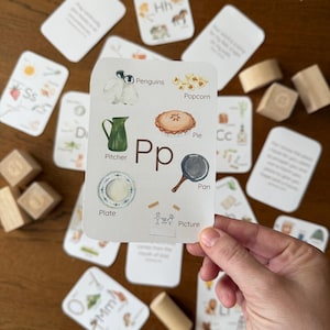 May include: A collection of educational flashcards featuring watercolour illustrations of objects and animals, such as penguins, popcorn, and a pie. Wooden blocks and a cylinder are also present.