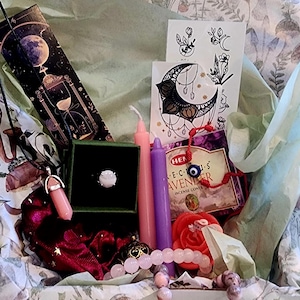 Puede incluir: Una caja de regalo llena de varios art&iacute;culos, incluyendo un anillo, velas, una pulsera, un collar, conos de incienso y tatuajes temporales. La caja est&aacute; forrada con papel de seda y papel decorativo. Los art&iacute;culos son en tonos rosa, morado y verde.