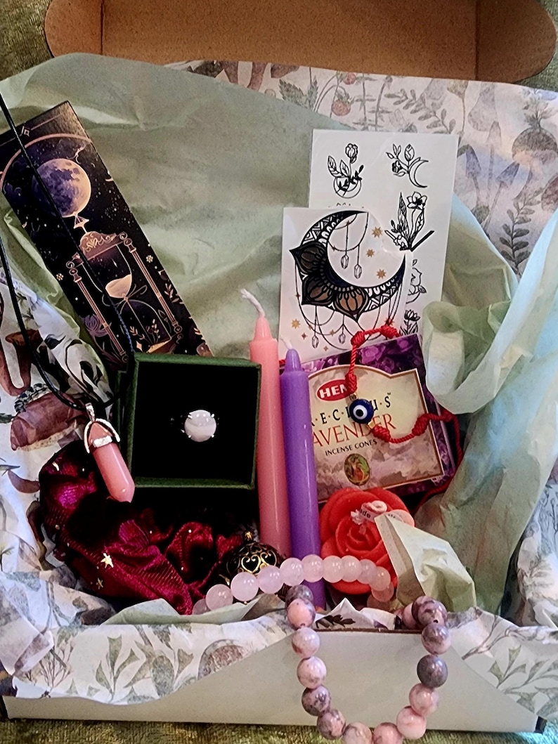 Puede incluir: Una caja de regalo llena de varios art&iacute;culos, incluyendo un anillo, velas, una pulsera, un collar, conos de incienso y tatuajes temporales. La caja est&aacute; forrada con papel de seda y papel decorativo. Los art&iacute;culos son en tonos rosa, morado y verde.