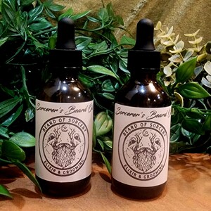 May include: Two dark glass bottles of beard oil with black droppers. Each bottle has a label with the text "Sorcerer's Beard Oil" and a circular graphic with a beard design. The words "Beard of Sorcery" and "Elixir & Grooming" are also on the label.