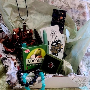 Puede incluir: Caja de regalo abierta con varios art&iacute;culos: collar, coletero, incienso, pulseras, anillo y tarjetas. La caja contiene una caja de incienso verde con el texto "Coconut Incense Co." y un anillo en una caja verde.
