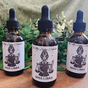 May include: Three brown glass bottles with black droppers. Each bottle has a white label with a drawing of a woman holding scales and the words "The Miss Zodiac Collection" and "MISS LIBRA". The bottles are on a wooden surface.