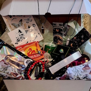 Puede incluir: Una caja blanca abierta llena de varios art&iacute;culos espirituales. Incluye collares con colgantes de cristal, incienso, una vela, un anillo, cuentas, pegatinas y una varita de selenita. La caja est&aacute; forrada con papel de seda verde.