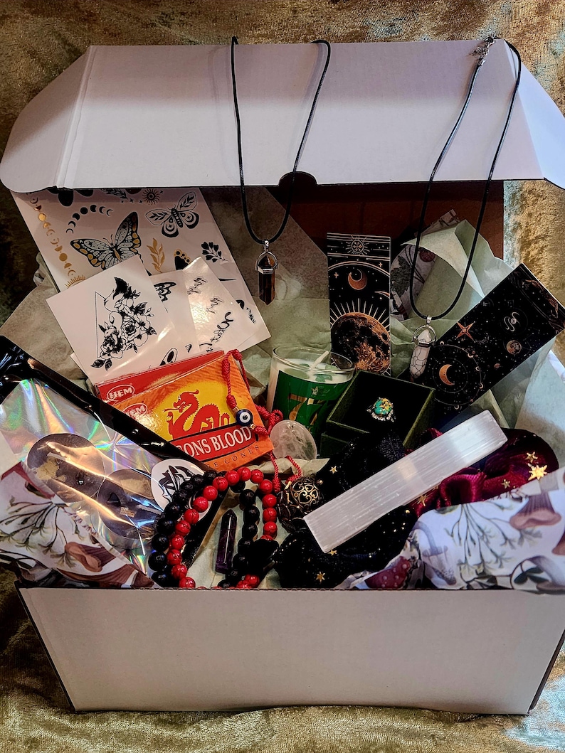 Puede incluir: Una caja blanca abierta llena de varios art&iacute;culos espirituales. Incluye collares con colgantes de cristal, incienso, una vela, un anillo, cuentas, pegatinas y una varita de selenita. La caja est&aacute; forrada con papel de seda verde.