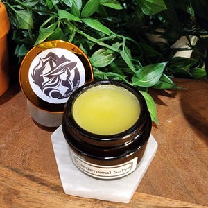 Puede incluir: Un pequeño frasco de vidrio ámbar oscuro lleno de un ungüento amarillo semisólido. El frasco tiene una etiqueta blanca con el texto Goldenseal Salve. La tapa es de oro metálico con un logotipo circular que muestra una figura de zorro.
