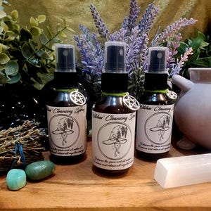 Puede incluir: Tres botellas de spray de vidrio oscuro con tapas negras y etiquetas que dicen "Witches' Cleansing Spray". Cada botella tiene un dije de pentáculo plateado. Las botellas están rodeadas de vegetación, lavanda y cristales.