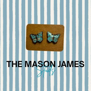 May include: A pair of butterfly-shaped stud earrings featuring a light blue and white floral pattern on a brown display card. The text THE MASON JAMES STUDS is printed below the earrings on a background of vertical light blue and white stripes.