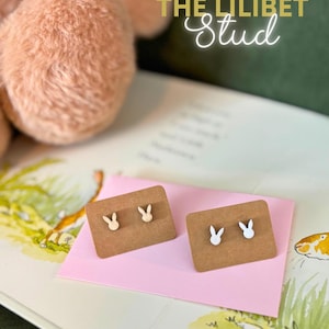 May include: Two pairs of bunny stud earrings on brown cards, one pair is light wood, the other is white. The earrings are displayed on a pink envelope, with a plush toy and a book in the background. The text "THE LILIBET Stud" is in the background.