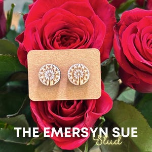 May include: Wooden stud earrings with a dandelion design, displayed on a brown card, set against a backdrop of vibrant red roses. The text "THE EMERSYN SUE Stud" is visible at the bottom of the image. These earrings are a perfect accessory for any outfit.