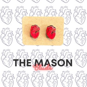 May include: A pair of red heart-shaped earrings with gold accents, displayed on a tan card. The background features a repeating pattern of anatomical heart illustrations. The text "THE MASON Studs" is at the bottom.