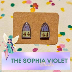 May include: A pair of stud earrings with a church window design. The earrings are purple and blue with the number 44 in a green box. The earrings are on a brown card with the text "THE SOPHIA VIOLET Studs".