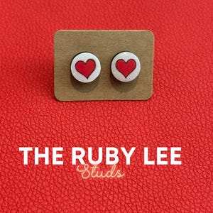 May include: A pair of heart-shaped stud earrings. Each earring features a red heart design on a white background, set on a small, rectangular cardboard display. The text "THE RUBY LEE Studs" is displayed in white.