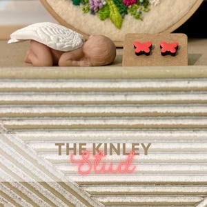 May include: A small sculpture of a baby with white angel wings, and a pair of red butterfly stud earrings on a brown card. The text "THE KINLEY Stud" is displayed in pink neon.